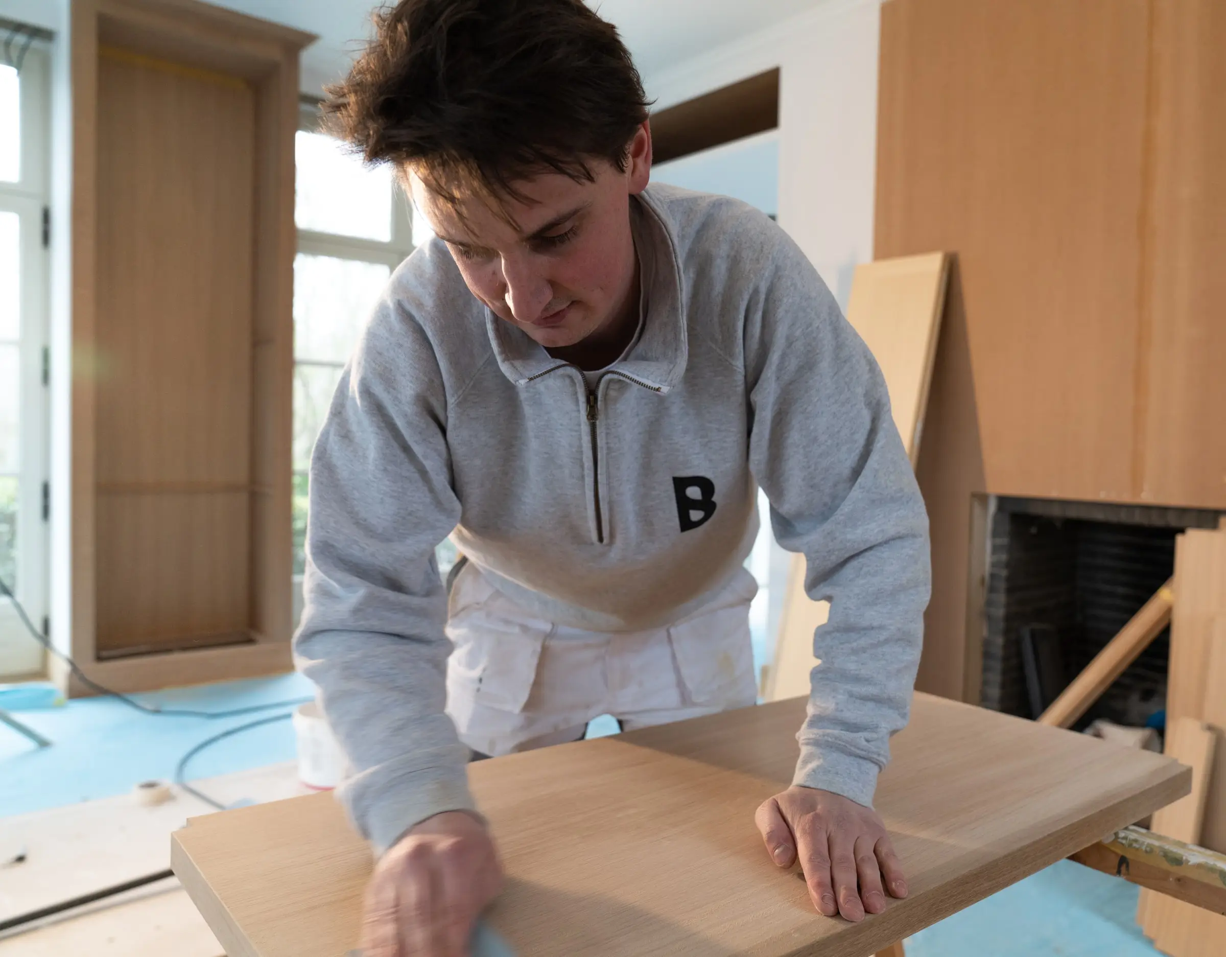 Timothy Blondeel schuurt een houten paneel op een werktafel, in een grijze trui met het logo