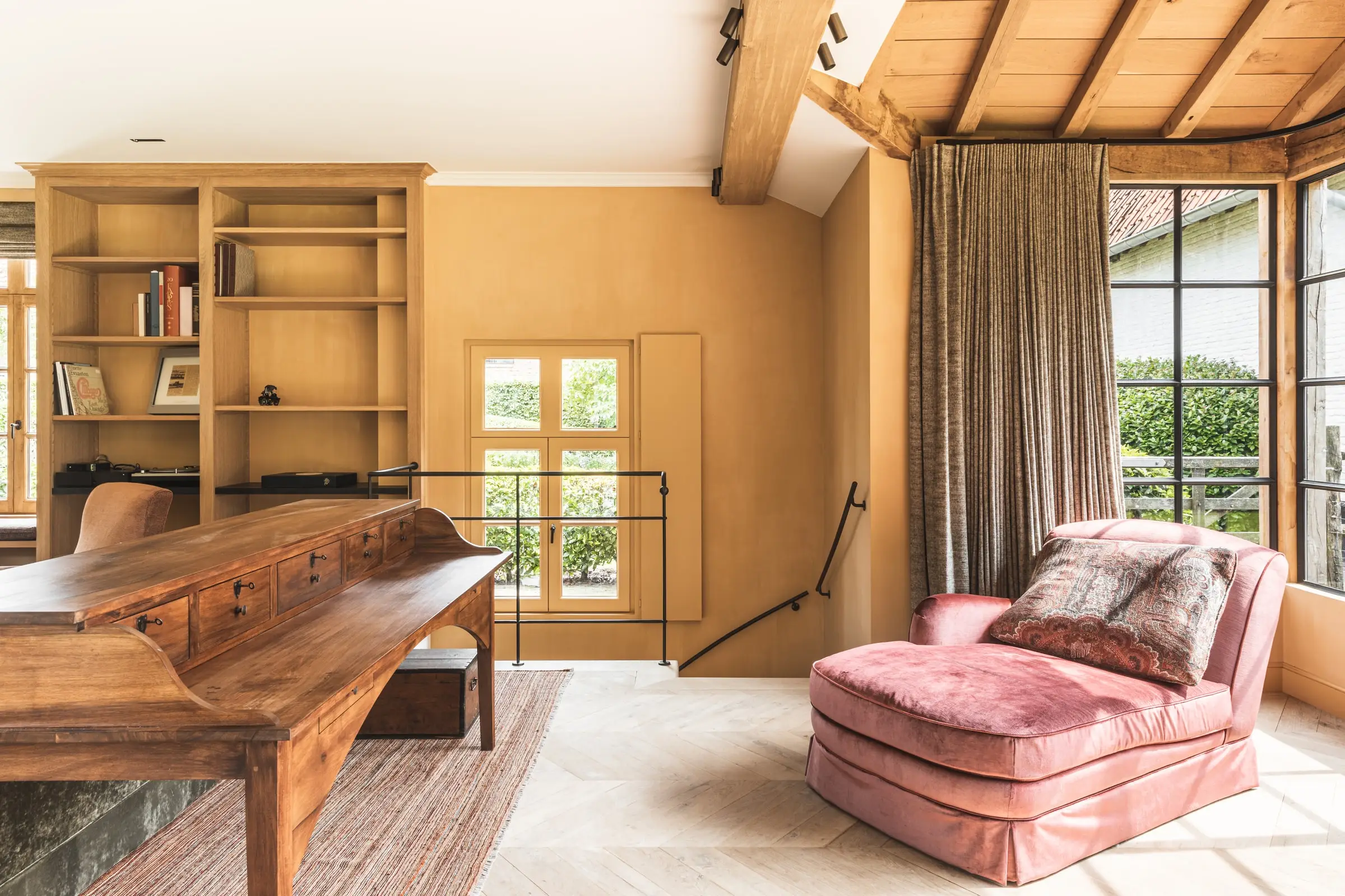 Woonkamer met een houten bureau, een roze chaise longue en zicht op de serre